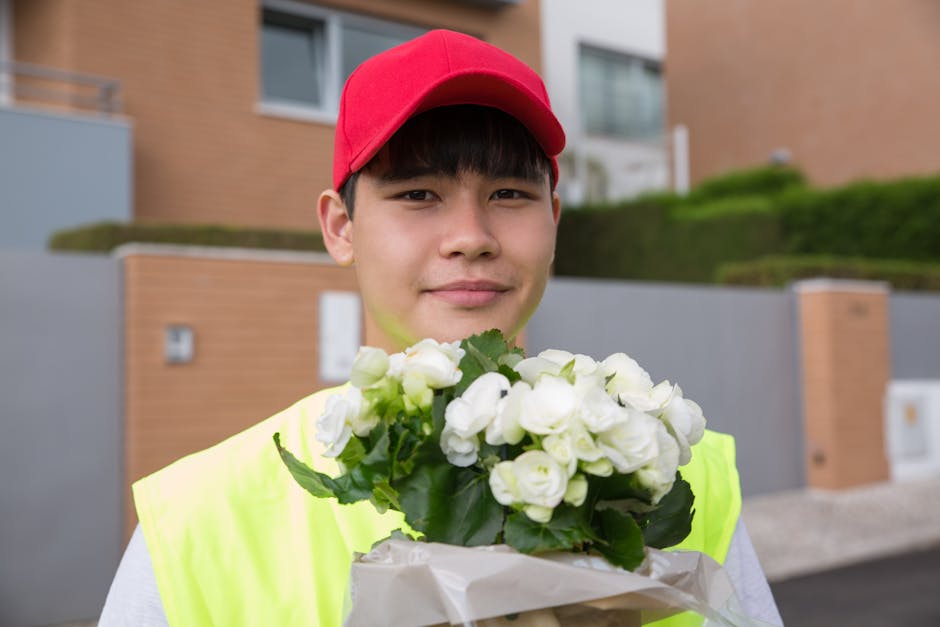 A young delivery person wearing a red cap and a bright yellow high-visibility vest is standing outdoors in a residential area with modern brick and concrete houses, holding a bouquet of fresh white fl