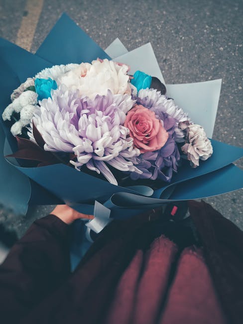 A vibrant bouquet featuring a mix of fresh flowers, including a pink spray rose, soft purple and white chrysanthemums, accented with small blue flowers, all arranged in a lush, full style. The bouquet