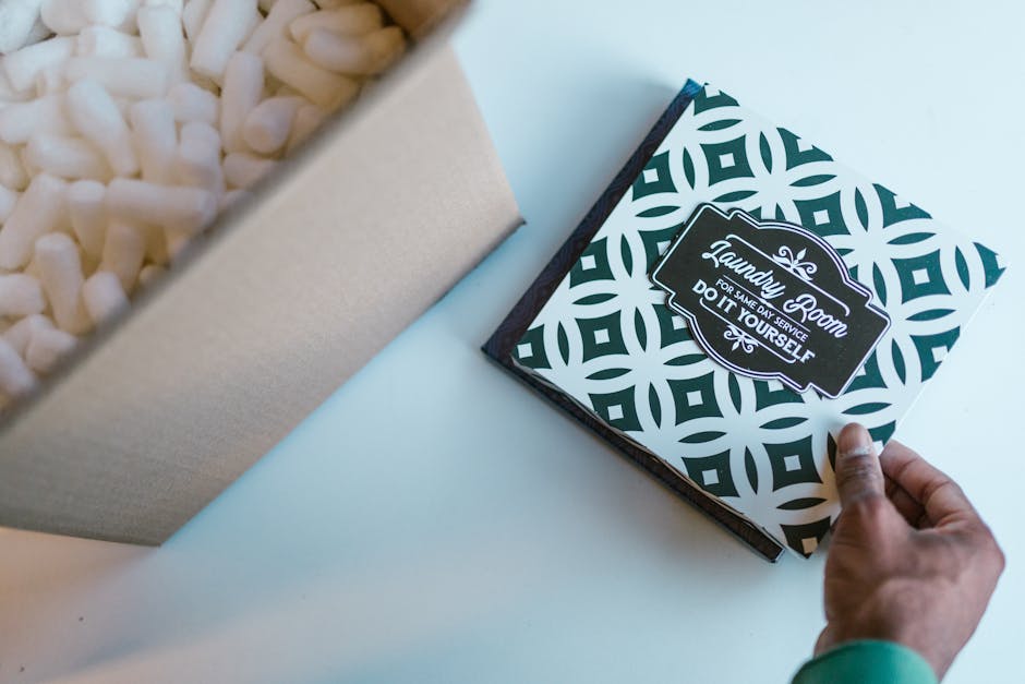 A close-up image of a person’s hand holding a decorative box with a geometric pattern and a label that reads 'Laundry Room for same day service, do it yourself,' positioned on a light-colored surfac