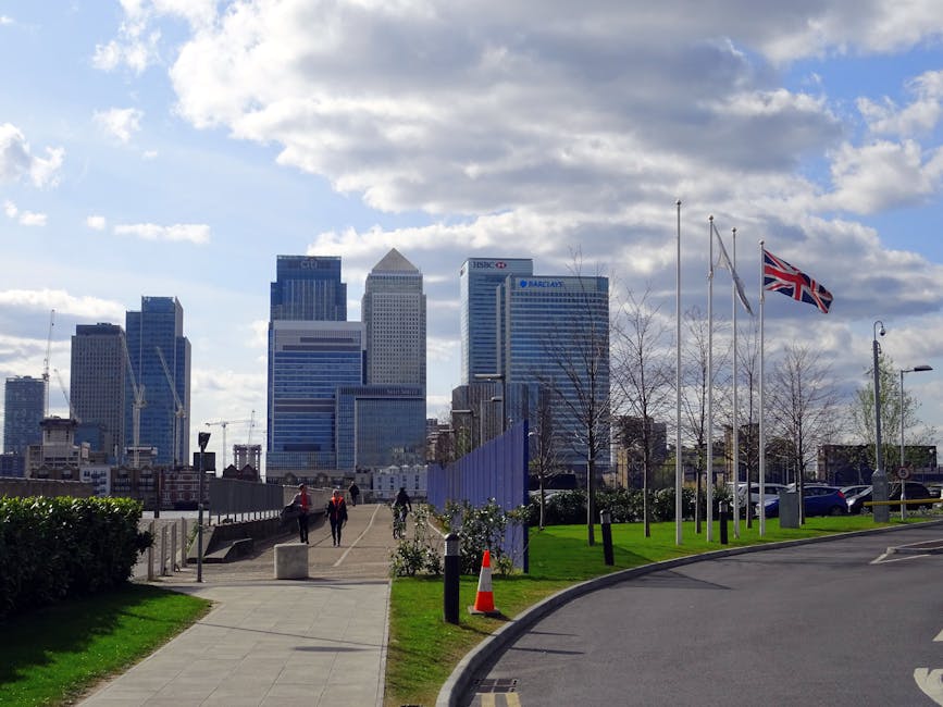 A vibrant bouquet of fresh flowers wrapped in white paper with a blue ribbon is being held by a person outdoors with a backdrop of the London skyline, including modern skyscrapers and flags on flagpol