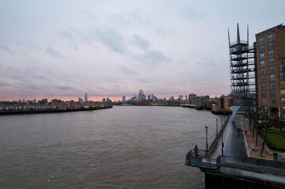 A view of the River Thames at dusk, with calm waters reflecting the soft pastel colors of the cloudy sky. On the right side, modern brick apartments and a steel scaffold-like structure are visible alo