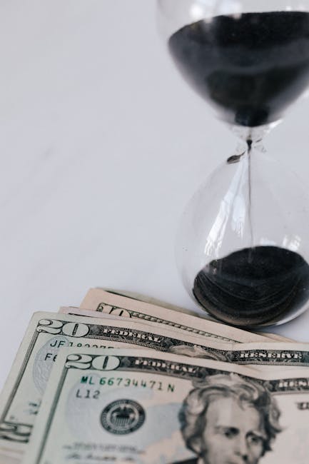 An hourglass with black sand positioned upright next to a stack of US twenty-dollar bills, placed on a white surface with a plain white background. The hourglass's glass bulbs are transparent, showcas