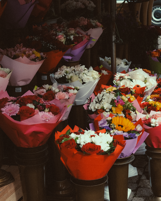 A selection of freshly arranged flower bouquets displayed on black tables outside a flower shop, featuring a variety of blooms such as red roses, white lilies, sunflowers, pink gerbera daisies, purple