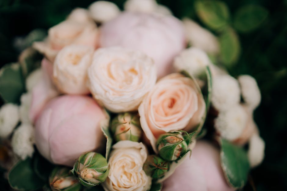 A close-up of a floral bouquet featuring soft peach and blush-colored roses with delicate, layered petals, mixed with small white blossoms and green buds. The flowers are arranged in a dense, rounded
