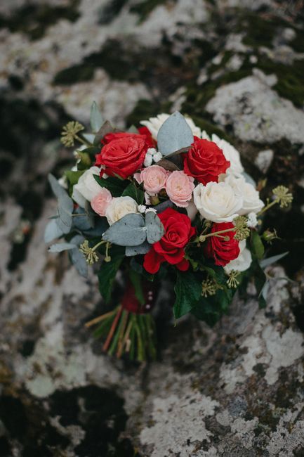A bouquet of fresh flowers featuring red and white roses, soft pink ranunculus, and sprigs of green foliage, accented with dusty blue eucalyptus leaves. The arrangement is beautifully compact, with th