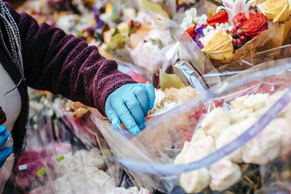 A person wearing a dark purple textured fleece and blue disposable gloves is selecting and handling a bouquet of fresh flowers wrapped in clear plastic in a flower shop. The arrangement includes roses
