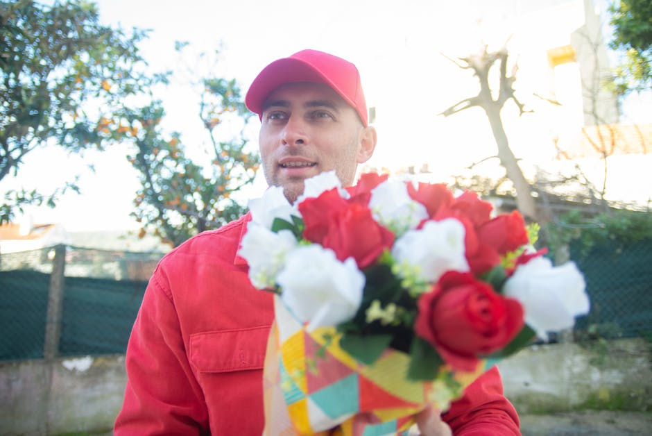 A male courier wearing a red cap and matching red jacket is delivering a floral gift outside on a bright day. He holds a bouquet of fresh red and white roses wrapped in colorful paper, with detailed p