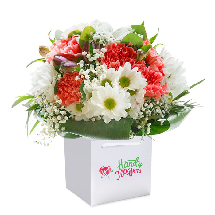 A vibrant, expertly constructed bouquet stands upright in a pristine square white gift bag, framed against a faultlessly white background to spotlight its beauty. Featuring a stunning contrast of hues, the arrangement melds lush, frilled carnations in shades of coral, peach, and bright red, their textured petals cascading in delicate layers. Interspersed are crisp white daisies, their golden-green centers reminiscent of sunlight glimmering over Old Ford's communal gardens, imparting a sense of optimism and charm. Finer details reveal dainty clouds of baby's breath scattered sparingly throughout-infusing a light, airy presence and a subtle sense of innocence. Look closely and you may notice hints of closed, purple-tinged buds tucked into the left side, adding complexity and suggesting gentle anticipation. Encircling the display, deep green, glossy leaves form a stately collar, contributing substance and depth to the bouquet-as if drawing inspiration from the local parklands bordering the Hertford Union Canal. The white gift bag, designed with sturdy white handles for ease of carrying through the busy streets of Old Ford, highlights the arrangement's readiness for gifting on any special occasion. Beautifully composed, the bouquet's rounded form and fresh, vibrant palette convey celebratory promise-be it for a birthday, an anniversary, or a simple gesture of kindness delivered locally. This image eloquently presents the care and expertise with which Old Ford's florists approach every creation, capturing both the artistry and heartfelt intention behind every stem.