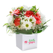 There is a gentle exuberance radiating from this lush bouquet, artfully nested within a flawless white gift bag-its sturdy ribbon handles inviting you to deliver a bit of joy along the leafy avenues of Old Ford. The central flourish of soft coral and vivid red carnations, each petal frilled with generous texture, blends harmoniously with clusters of snowy daisies sporting golden-green cores, reminiscent of wildflowers found near the edges of the Olympic Park. Wisps of white baby's breath float delicately between, imparting an ethereal softness that calls to mind a refreshing morning breeze along Roman Road. Touches of purple-tinged buds peek subtly from the left side, their quiet presence adding notes of intrigue and heralding blossoms yet to open. All of this is encircled by a rich mantle of deep green foliage-some leaves broad and gleaming, others more slender-giving the arrangement both body and elegance. The bouquet forms a rounded, poised silhouette, exuding abundance and fresh vibrancy from every angle. Illuminated by soft, even lighting, the composition is visually uplifting and pristine, echoing the freshness of a local florist's morning selection. Whether intended as a heartfelt gift to celebrate a birthday, anniversary, or simply wish a neighbour well, this carefully-crafted arrangement perfectly captures the spirit of community and thoughtful giving found throughout Old Ford.